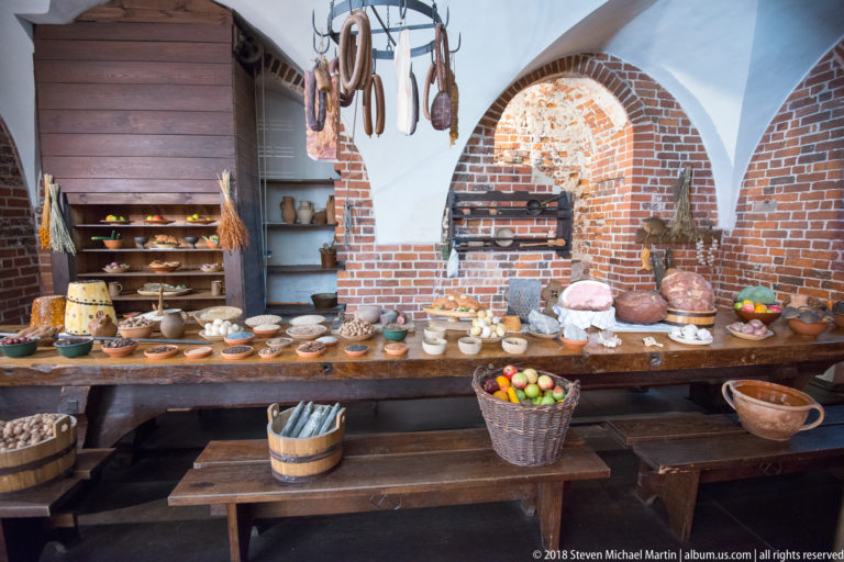 Long wooden kitchen table covered with bowls, pottery, baskets, utensils, and food, with hanging sausages and shelves under brick arches.
