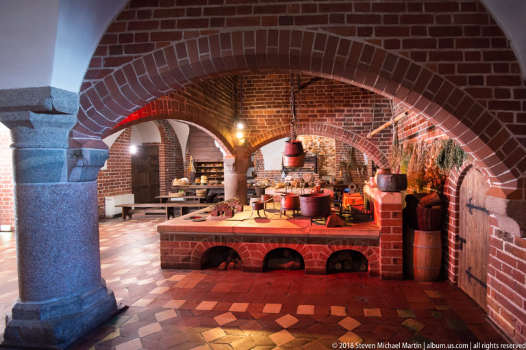 Brick-vaulted castle kitchen with raised cooking hearth, pots, kettles, tiled floor, and heavy masonry arches.
