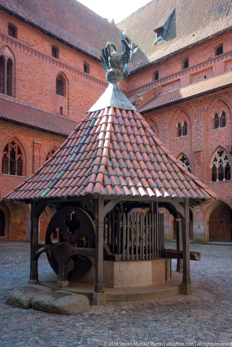 Roofed timber courtyard well with a large winding wheel and dragon finial, surrounded by red-brick Gothic façades.