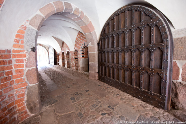 Large iron-studded wooden gate set in a brick-and-stone vaulted passage with cobbled floor and repeating arches.