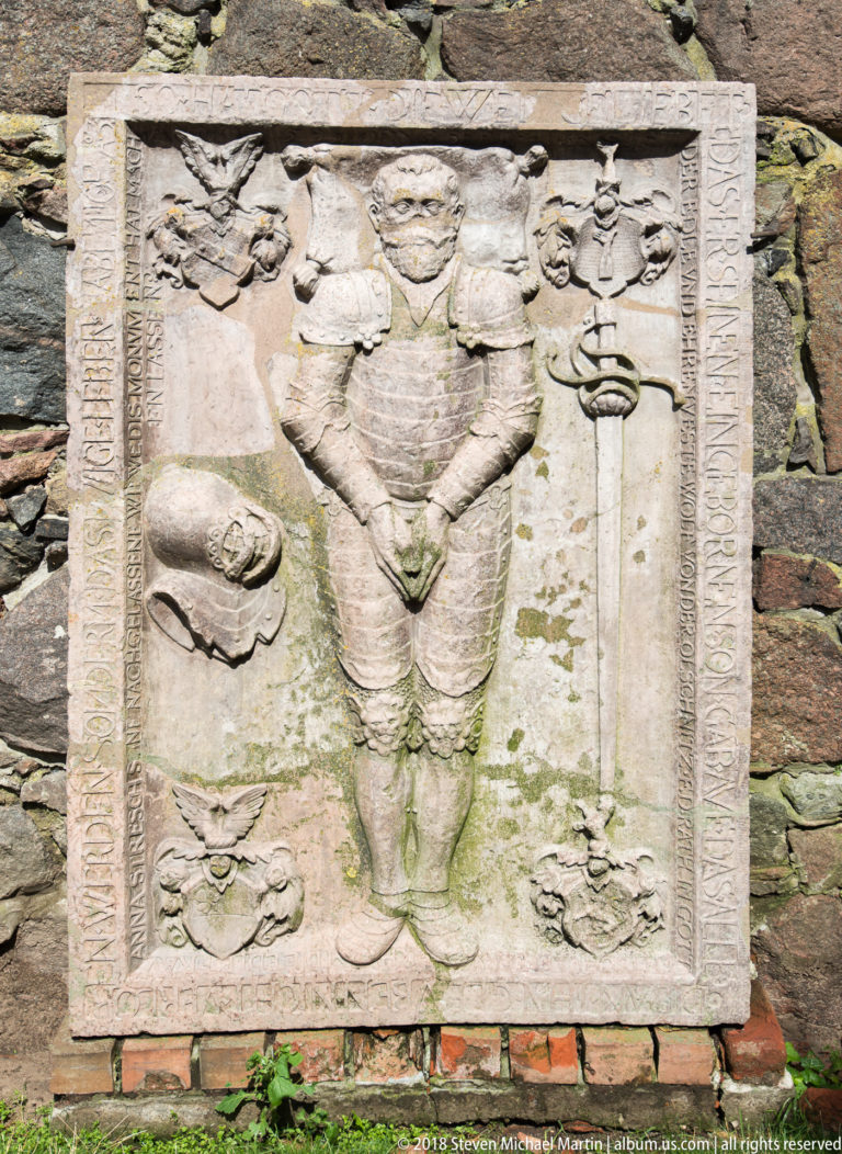 Weathered rectangular stone relief of a standing man in period dress, flanked by heraldic or symbolic emblems.