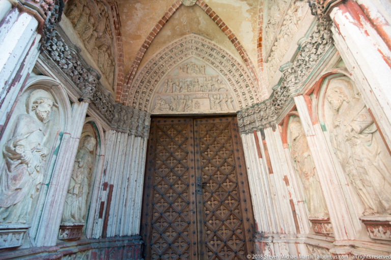 Frontal view of a Gothic portal with carved narrative tympanum, layered archivolts, niche figures, and patterned double doors.