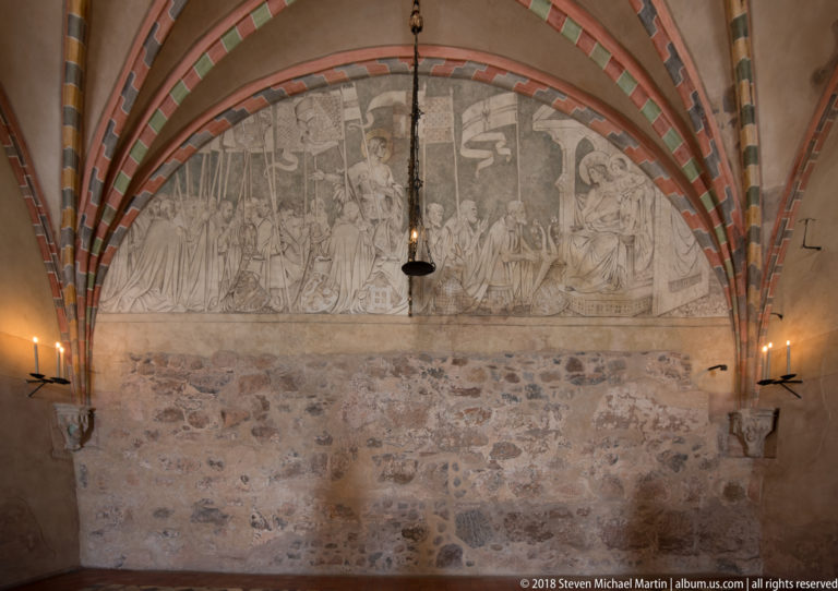 Large faded monochrome mural of many figures beneath striped Gothic vault ribs, with wall candles and a hanging lamp.