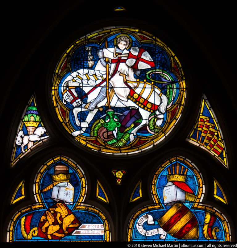 Stained-glass window with Saint George on horseback attacking a green dragon above smaller heraldic roundels.