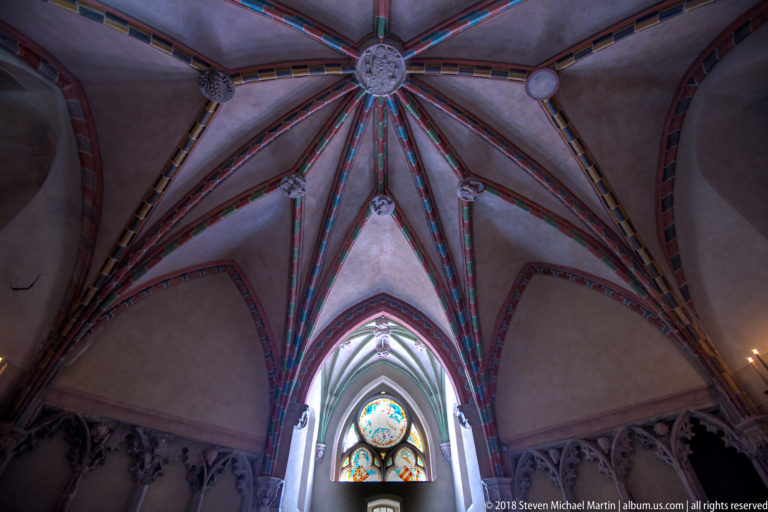 Painted Gothic rib vault with a central carved boss, multicolored ribs, and a stained-glass window below.