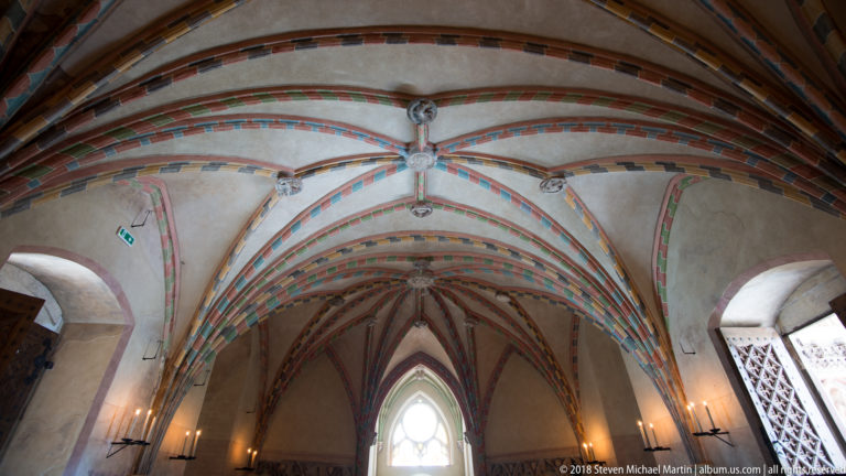 Wide interior view of painted Gothic rib vaults converging toward a tall pointed window, with carved bosses and wall sconces.