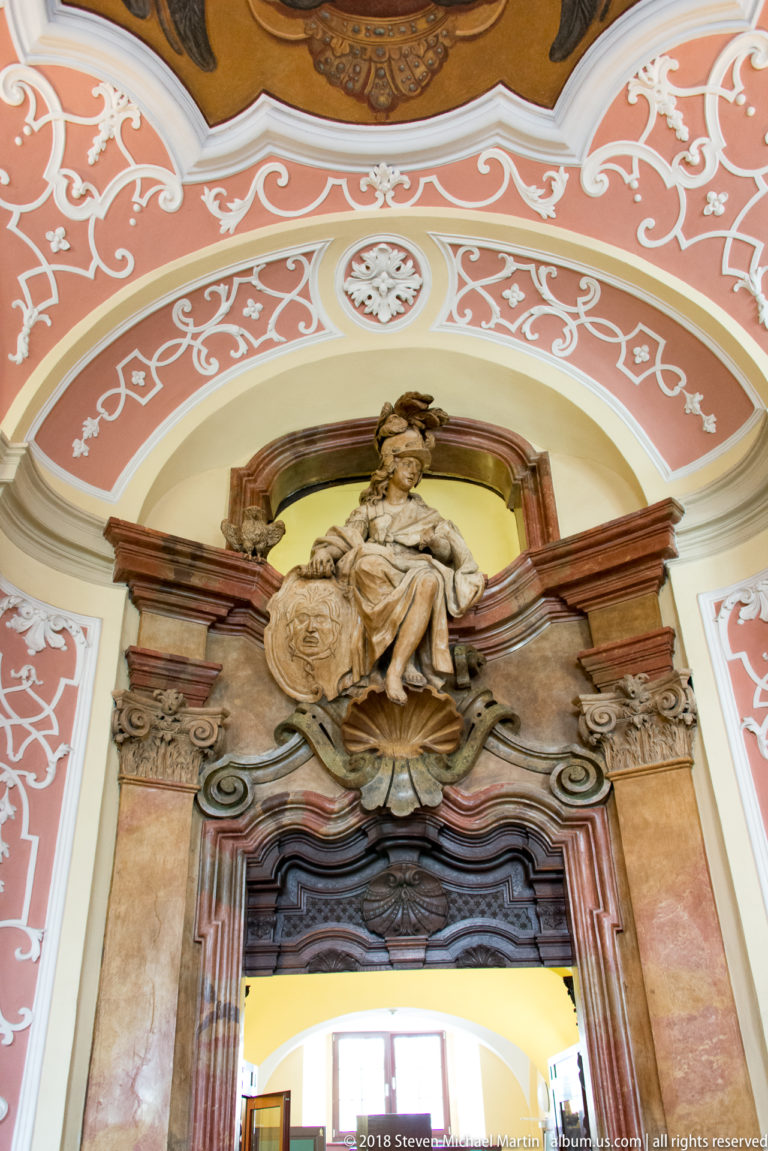 Baroque sculptural overdoor with seated female figure, child, shield, dark wood surround, and pink-and-white stucco decoration.