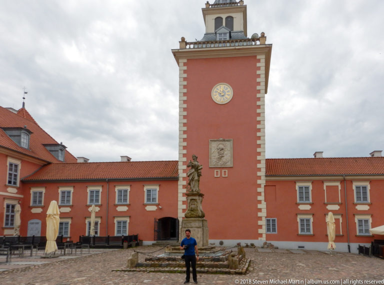 Pink palace range with tall clock tower, pale quoins, rooftop balustrade, stone monument, and cobbled courtyard.