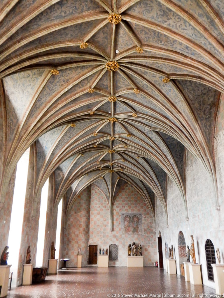 Long high hall with elaborate rib vaults, tall windows, brick walls, and sculptures displayed on low plinths.