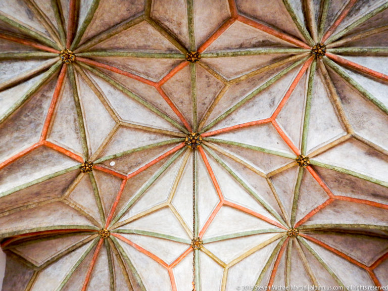 Close view of a star vault with multicolored painted ribs and gilded bosses against pale plaster.