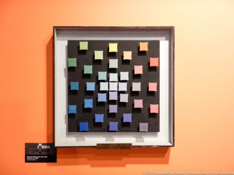 Framed abstract artwork of many small colored rectangles scattered across a black square field on an orange wall.