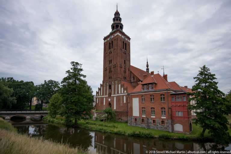 Wide view of a tall red-brick tower, adjoining buildings, bridge, trees, and reflective water.