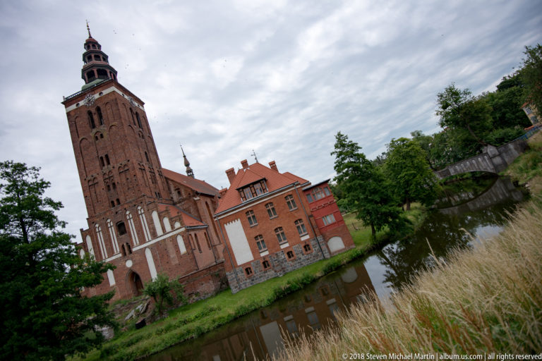 Tall red-brick Gothic tower and adjoining historic buildings beside a calm waterway.