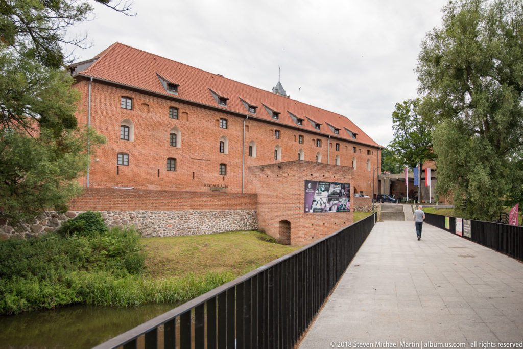 Long red-brick castle building beside a pedestrian bridge, grassy bank, mature trees, and masonry base.