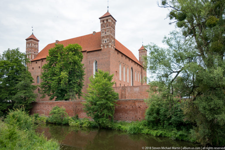 Large red-brick castle with corner towers and steep tiled roofs rising above water and a brick retaining wall.