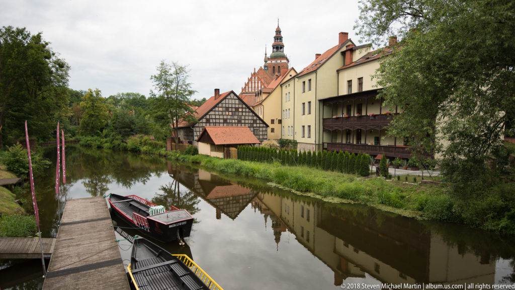 Small boats at a landing on calm water beside historic buildings, trees, and a tall tower in the background.