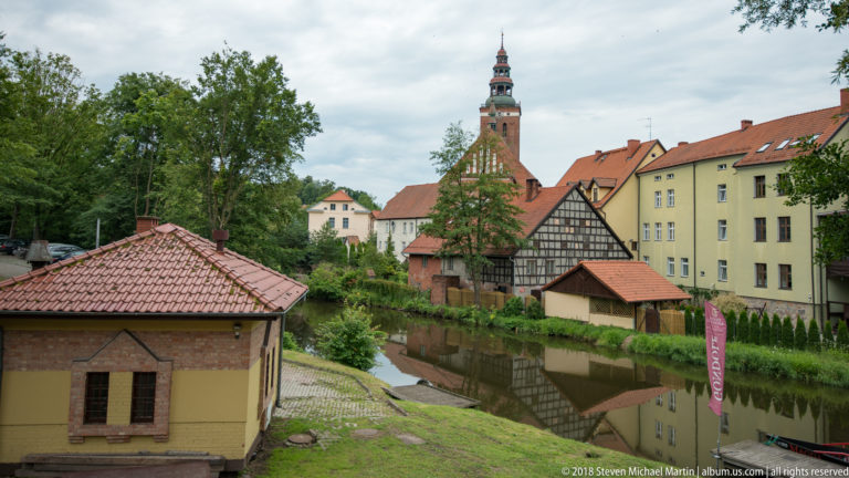 Narrow calm waterway beside half-timbered and plastered buildings, with trees and a tall distant tower.