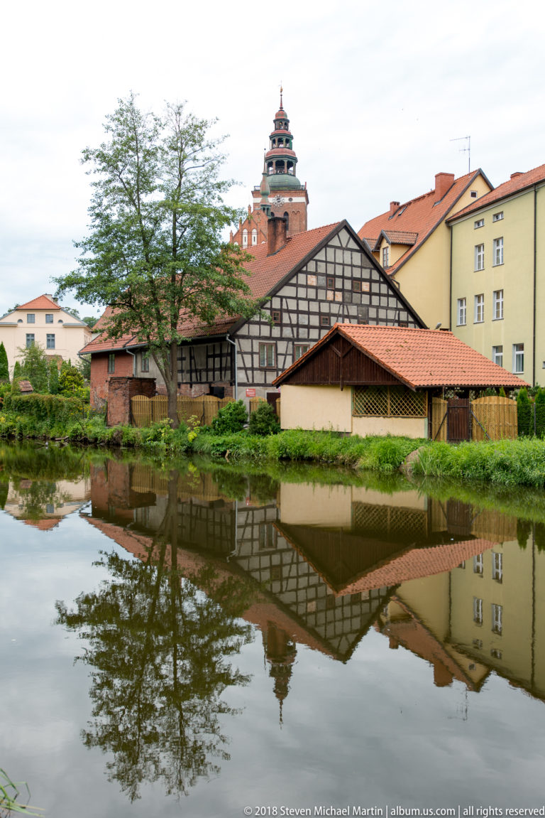 Half-timbered building and neighboring town houses reflected in calm water, with a tall tower rising in the distance.