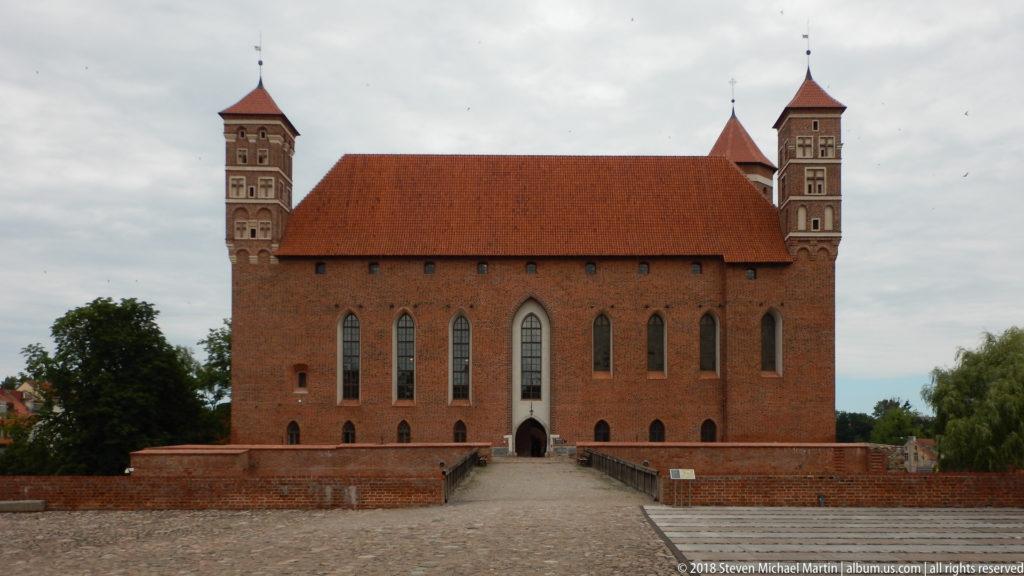 Frontal view of a large red-brick Gothic castle wing with pointed windows, steep tiled roof, and tall corner towers.