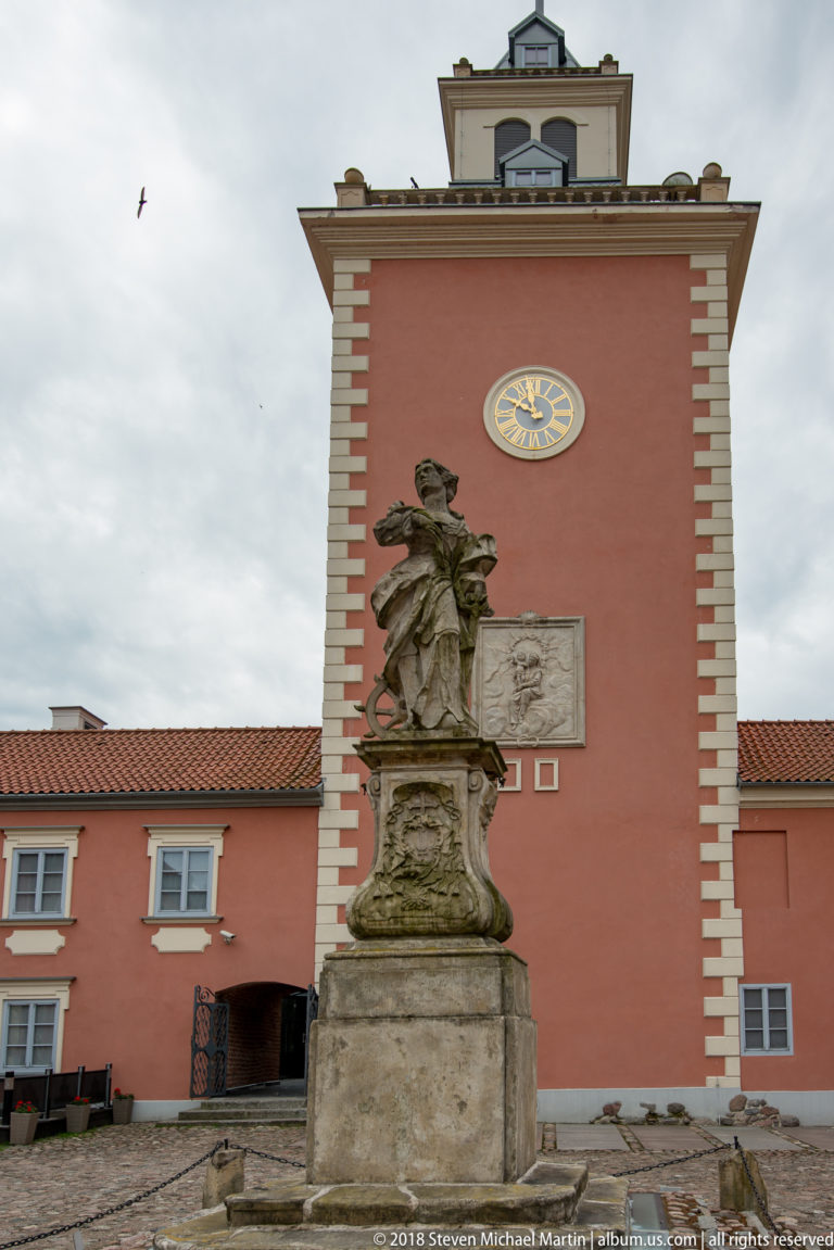 Pink palace tower with blue-and-gold clock, pale corner quoins, wall relief, and stone statue in front.