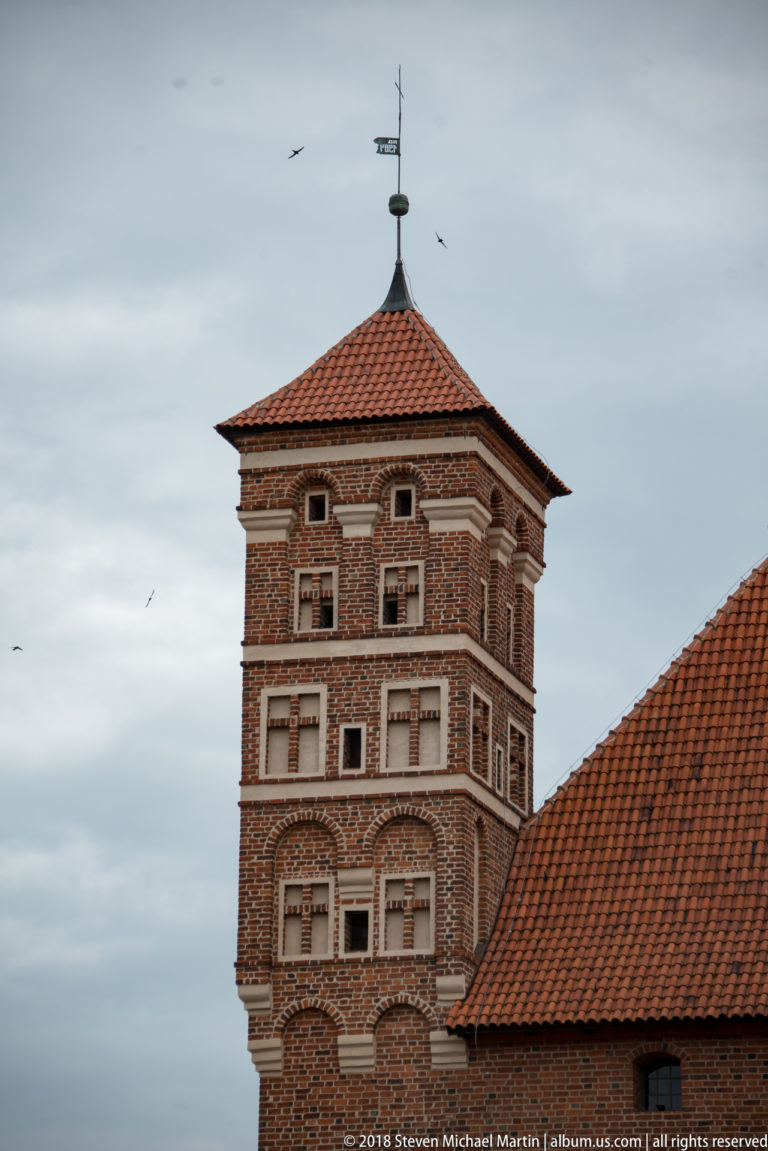 Square red-brick castle tower with pale decorative bands, small framed openings, and a red-tiled pyramidal roof.