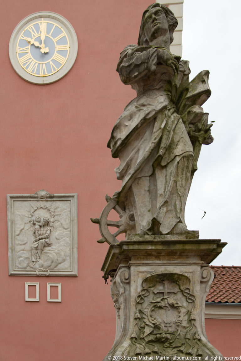 Weathered stone figure in flowing drapery on a carved pedestal before a pink wall with relief and clock.