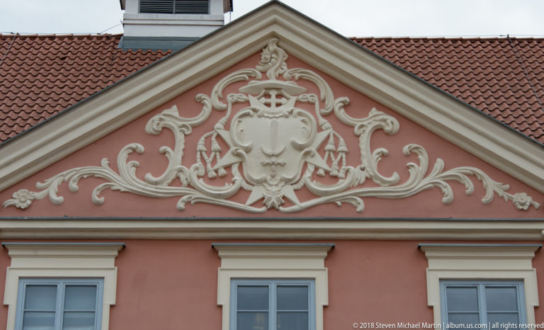 White stucco heraldic cartouche and scrolling ornament centered on the triangular pediment of a pink façade.