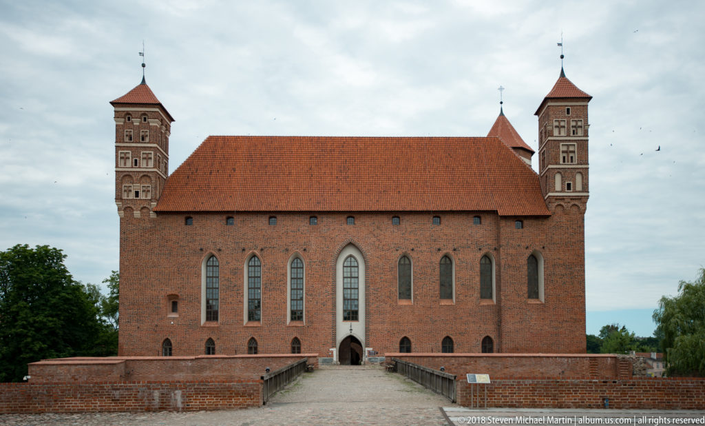 Centered view of red-brick castle with tall pointed windows, twin corner towers, tiled roof, and entrance at center.