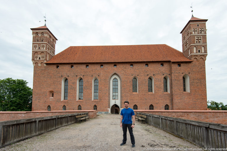 Large red-brick castle façade with tall Gothic windows, corner towers, red-tiled roof, and bridge approach.