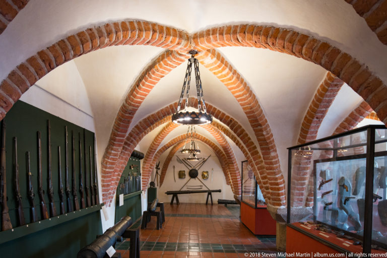 Long vaulted gallery with wall-mounted weapons, armor cases, chandeliers, and terracotta tile flooring.