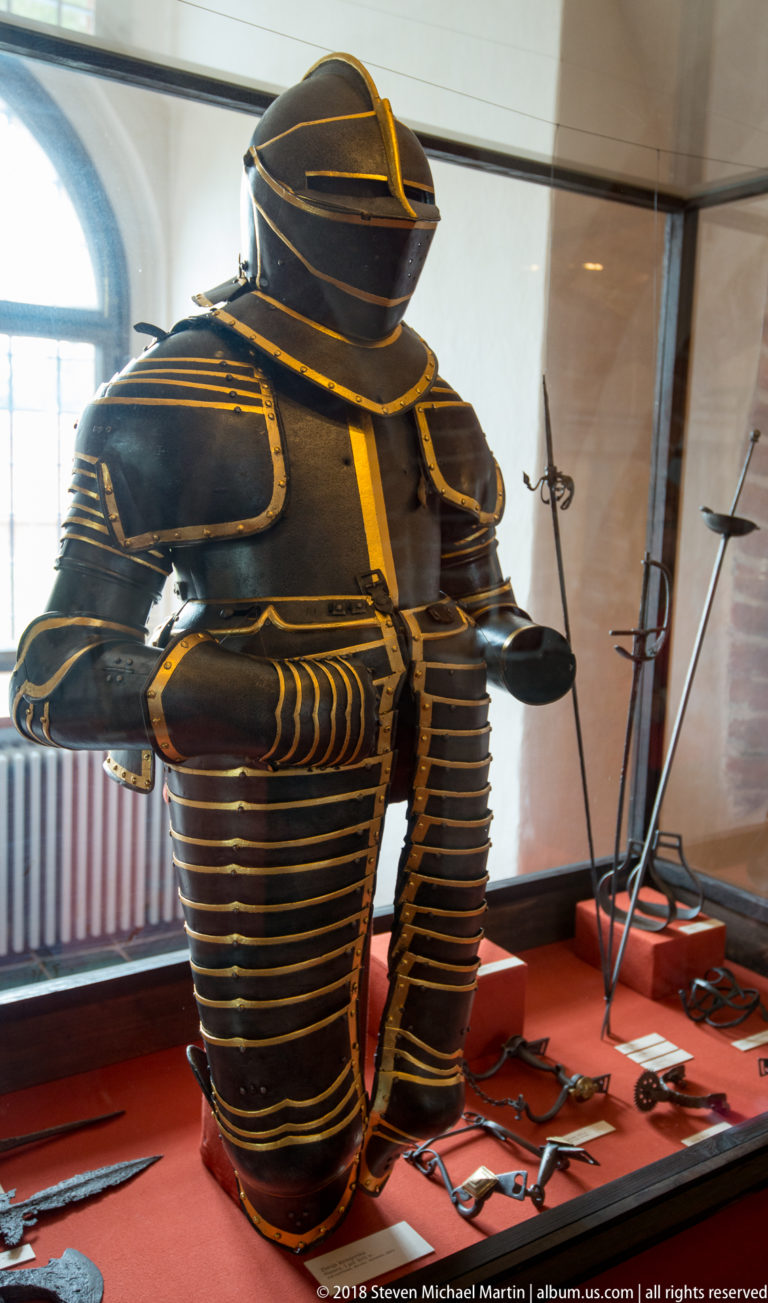 Full dark plate armor with gilt-edged bands displayed in a glass case among swords and polearms.