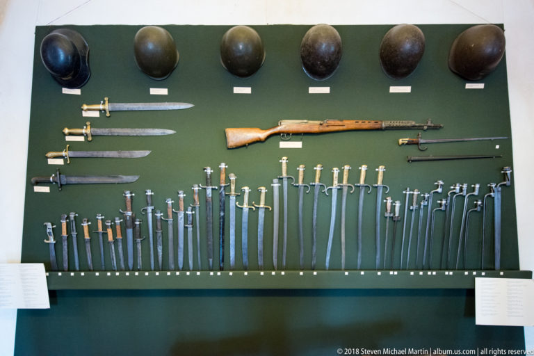 Large wall display of swords, sabers, daggers, bayonets, helmets, and a long firearm.
