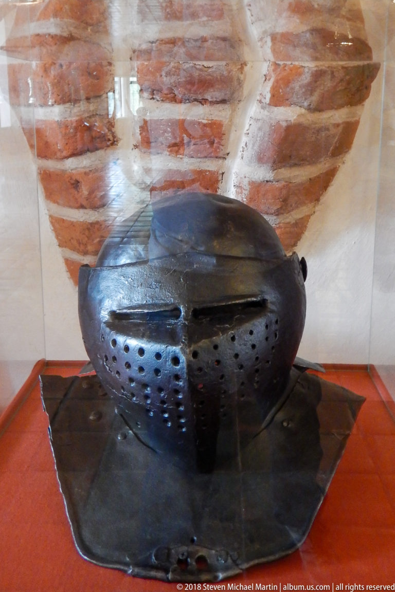 Dark iron helmet with perforated face plates and broad neck defense displayed against a brick-vaulted background.
