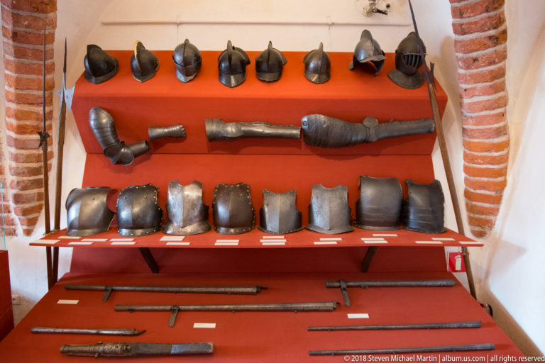 Historic helmets, arm defenses, breastplates, swords, and long weapons arranged on red museum shelving.