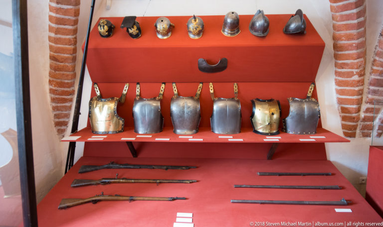 Red-backed museum display with helmets, breastplates, long firearms, and other historic weapons.