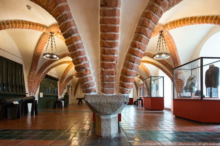Brick-vaulted gallery with weapon displays, armor cases, chandeliers, and a rough stone basin in the center.