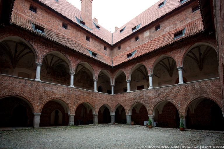 Enclosed cobbled courtyard surrounded by red-brick arcades, an upper columned gallery, and steep tiled roofs.