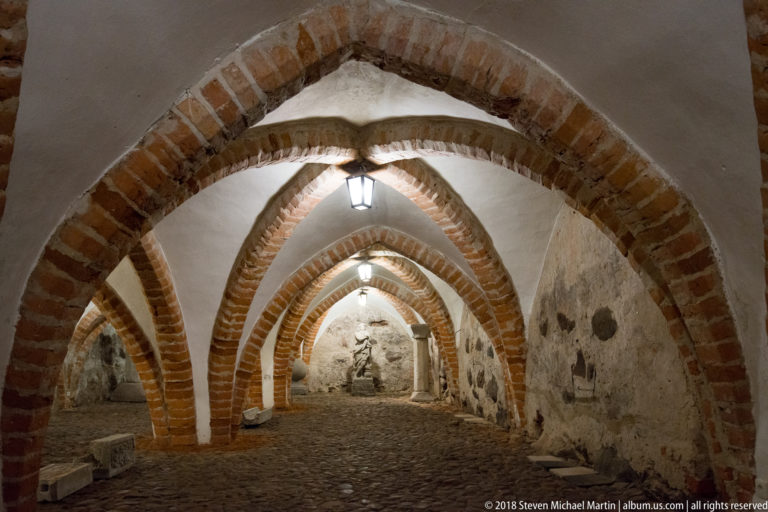 Deep view through repeated pointed brick arches in a dim stone-walled undercroft with cobbled floor and lanterns.
