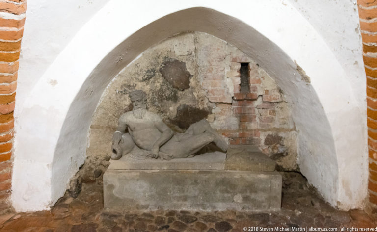 Weathered recumbent stone figure on a rectangular plinth beneath a whitewashed arch in a rough masonry interior.