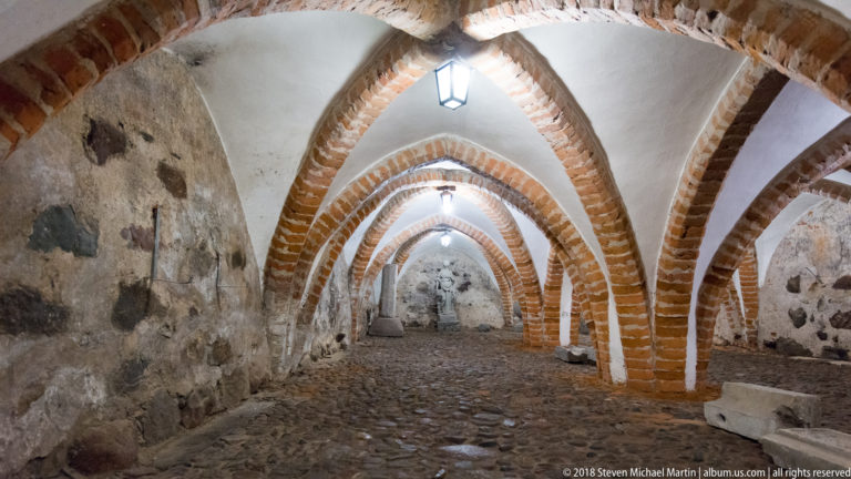 Low undercroft corridor with pointed red-brick arches, rough stone walls, cobbled floor, and hanging lanterns.