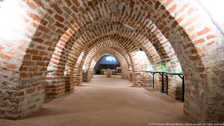 Long castle passage with repeated exposed-brick arches, thick masonry walls, modern lights, and museum cases.