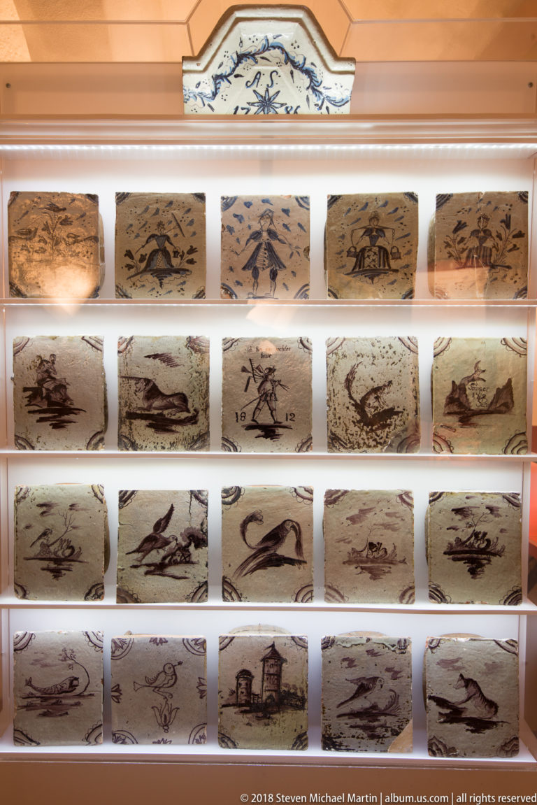 Rows of rectangular decorated ceramic tiles in a display case, each bearing a different animal, landscape, or architectural scene.