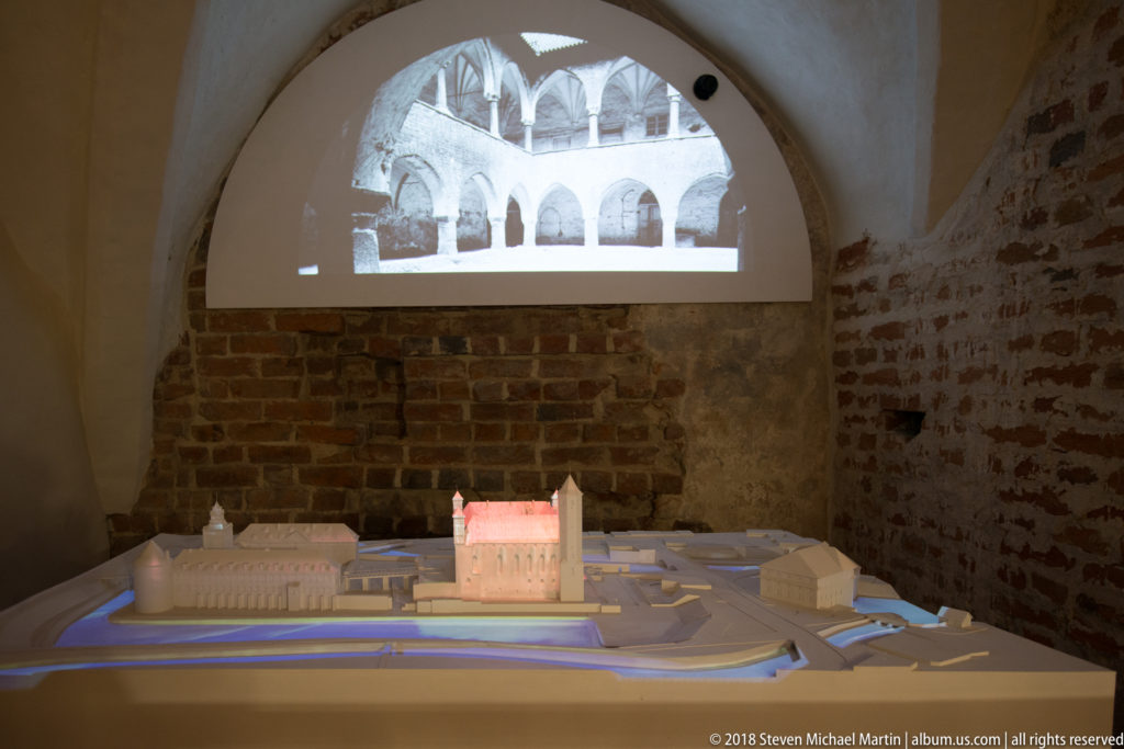Illuminated architectural model of a castle beneath a projected courtyard image in a dark brick-walled exhibit room.