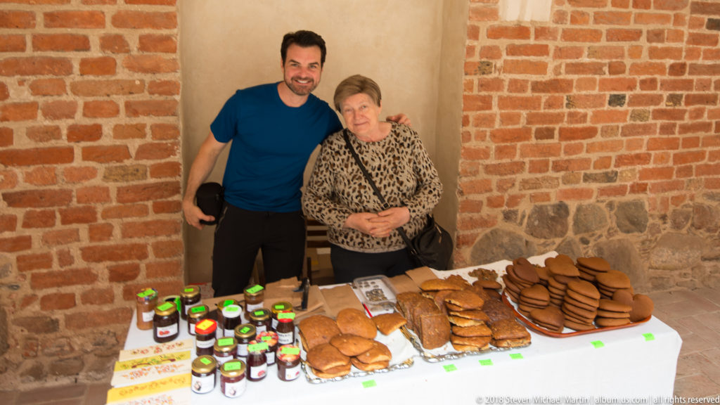 Two people pose behind a table of preserves, breads, cakes, and stacked heart-shaped gingerbread against brick walls.