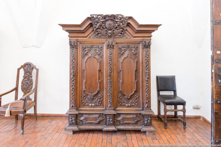 Large dark carved wooden cabinet with two paneled doors, ornate crest, relief scrollwork, drawers, and chairs beside it.