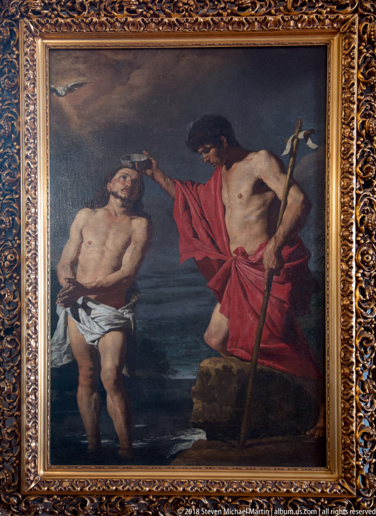 Framed religious painting showing John the Baptist in red beside Christ, with a long cross-staff against a dark background.