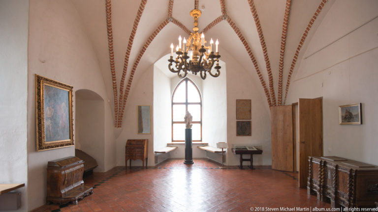 Narrow vaulted room with wooden chests, framed paintings, central chandelier, and a sculpture before a pointed window.