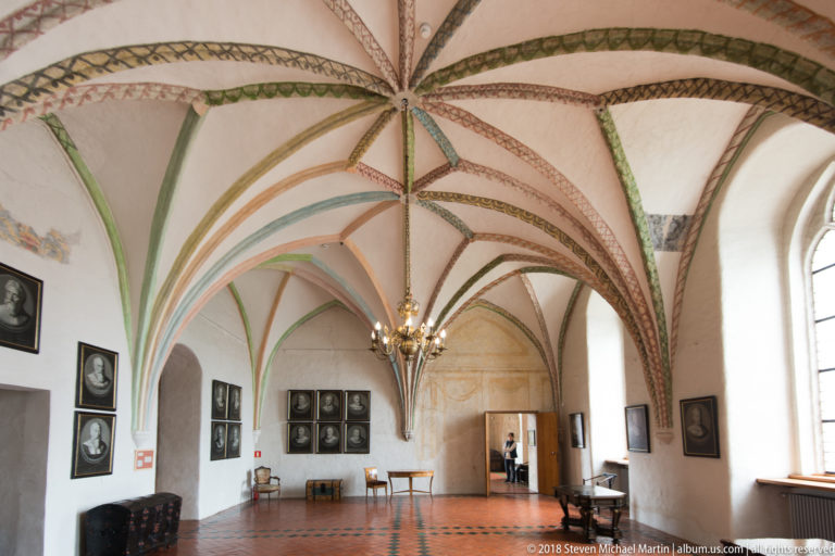 Wide vaulted room with painted ribs, portrait groups on the walls, red tile floor, chandeliers, and tall windows.