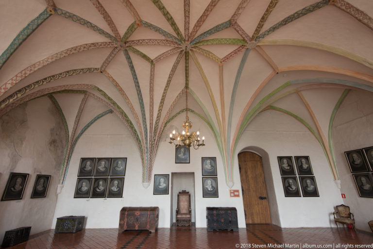 Vaulted white room with grouped black-framed portraits, wooden chests, a high-backed chair, and painted ceiling ribs.