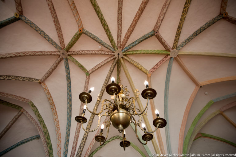 Brass chandelier suspended beneath a pale star vault with red, green, blue, and ochre painted ribs.